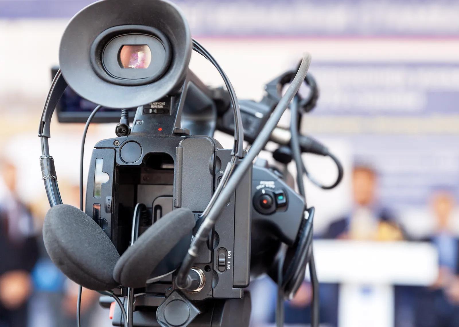 Fernsehkamera im Vordergrund, dahinter unscharf eine Person am Rednerpult